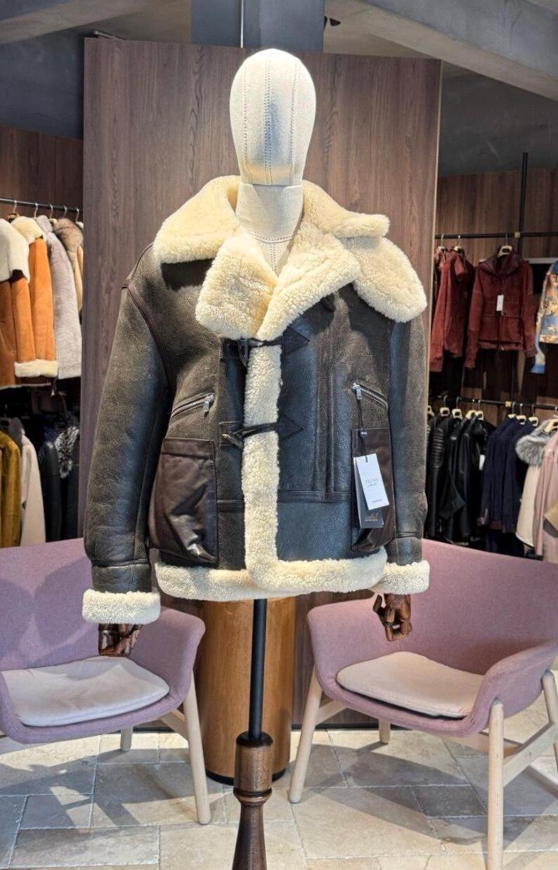 May include: A brown leather jacket with a cream-colored shearling lining and collar. The jacket features a front closure, two zippered pockets, and a large patch pocket. The jacket is displayed on a mannequin in a clothing store.