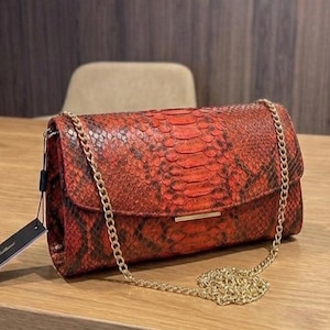 Könnte beinhalten: Eine rot-schwarze Handtasche mit Schlangenmuster und einem goldfarbenen Kettenriemen. Die rechteckige Tasche hat einen Klappverschluss und einen goldenen Balken. Ein schwarzes Etikett hängt an der Seite.
