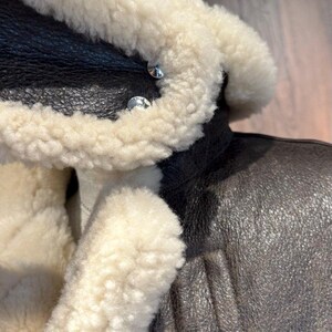 May include: Close-up of a brown leather jacket with a cream-colored shearling collar and lining. The jacket features silver-colored buttons and a textured leather exterior. A leather loop detail is visible.