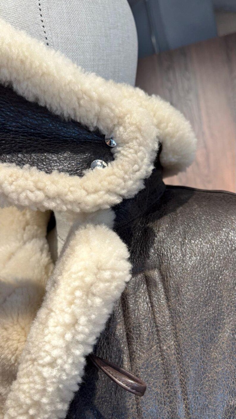 May include: Close-up of a brown leather jacket with a cream-colored shearling collar and lining. The jacket features silver-colored buttons and a textured leather exterior. A leather loop detail is visible.