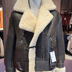 May include: A brown leather jacket with a cream-colored shearling lining and collar. The jacket features a double-breasted closure, two front pockets, and zippered chest pockets. A tag with the brand name is attached.