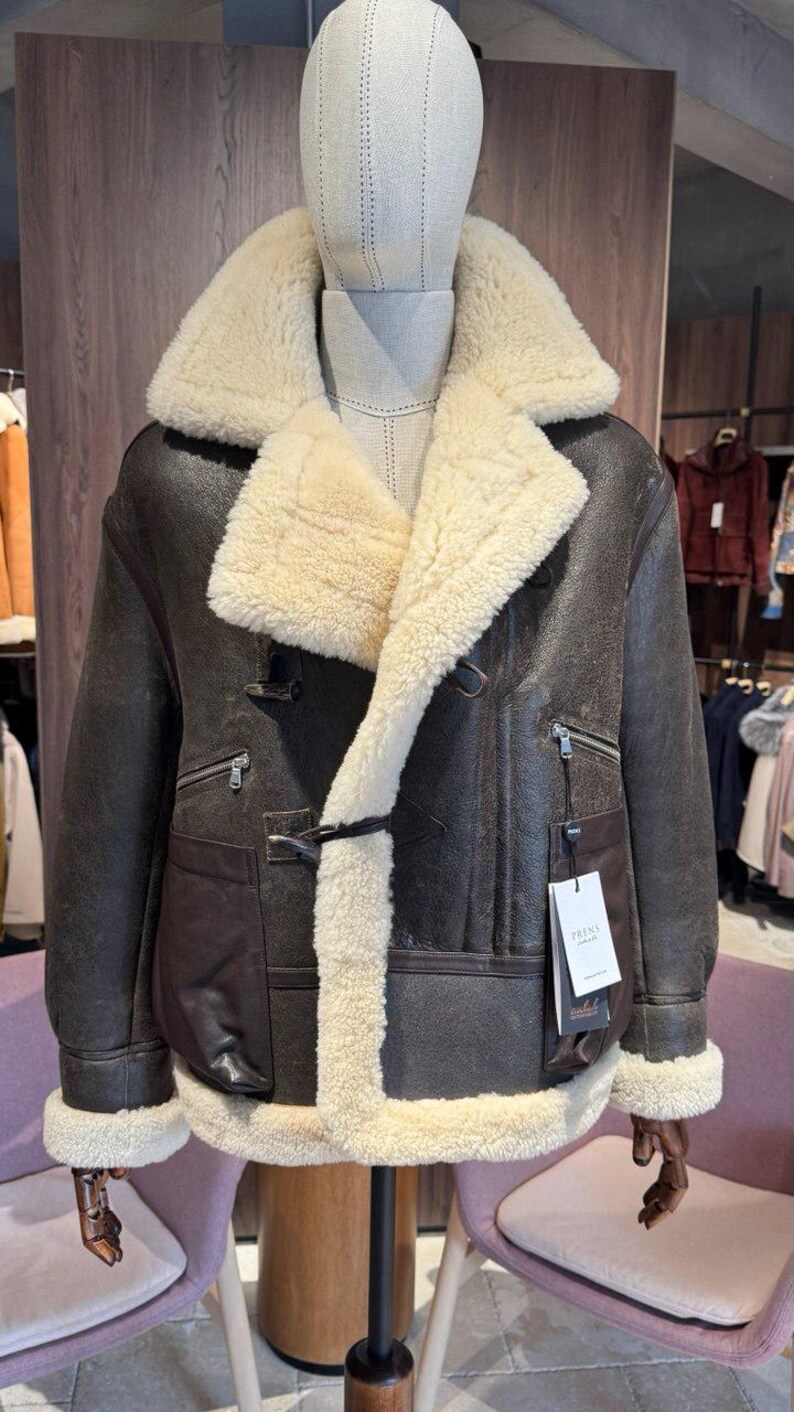 May include: A brown leather jacket with a cream-colored shearling lining and collar. The jacket features a double-breasted closure, two front pockets, and zippered chest pockets. A tag with the brand name is attached.