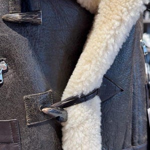 May include: Close-up of a brown leather jacket with a cream-colored shearling lining. The jacket features a toggle closure and a textured leather finish. The shearling trim is visible along the collar and front.