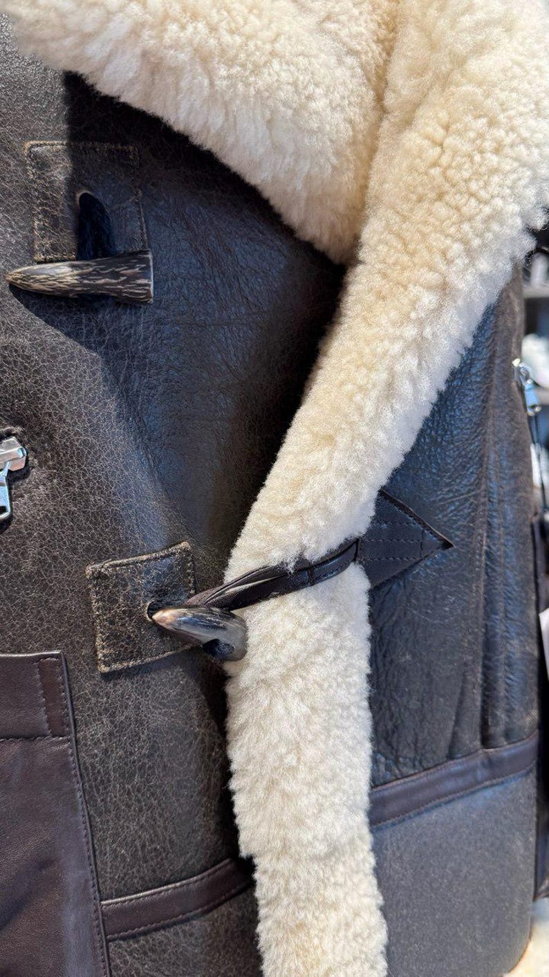 May include: Close-up of a brown leather jacket with a cream-colored shearling lining. The jacket features a toggle closure and a textured leather finish. The shearling trim is visible along the collar and front.
