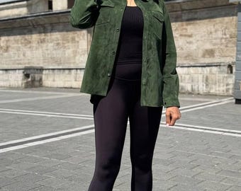 May include: A woman wearing a dark green suede shirt, black leggings, and black boots. She is also wearing black sunglasses. The outfit is suitable for casual wear. The background features a stone building.