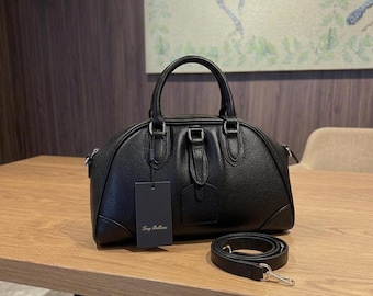 Puede incluir: Bolso de mano de cuero negro con asa superior y correa para el hombro desmontable. El bolso tiene forma redondeada, herrajes plateados y una etiqueta que dice "Tony Bellani". El bolso está sobre una superficie de madera.