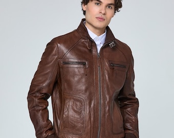 May include: A brown leather jacket with a zip-up front, two chest pockets, and two side pockets. The jacket has a ribbed hem and cuffs. The model is wearing a light blue shirt underneath the jacket.
