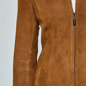 May include: A brown suede jacket with a full-length zipper closure. The jacket features a tailored fit with seams and stitching details. The sleeves have stitched elbow patches. The jacket is a warm, neutral color.