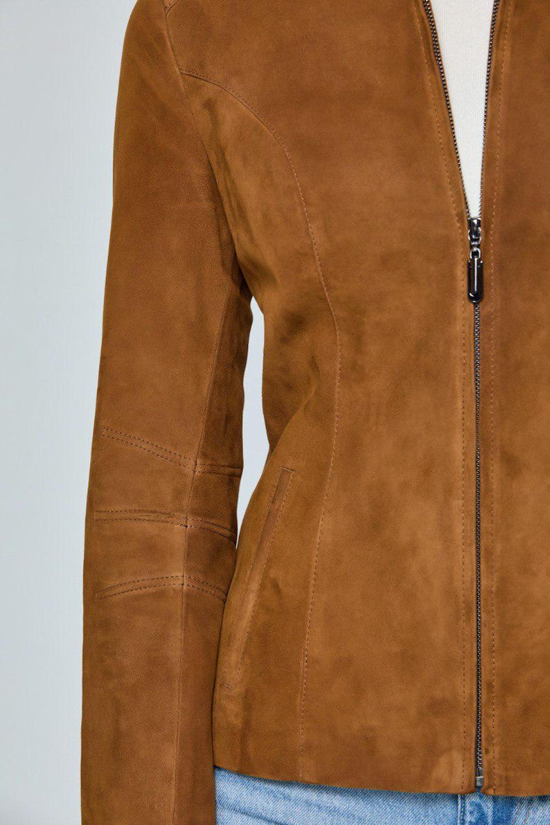May include: A brown suede jacket with a full-length zipper closure. The jacket features a tailored fit with seams and stitching details. The sleeves have stitched elbow patches. The jacket is a warm, neutral color.