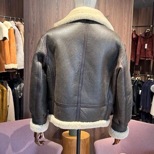 May include: A brown leather jacket with a cream-colored shearling collar and cuffs is displayed on a mannequin. The jacket has a classic design with a slightly distressed appearance. The back view shows the jacket's details, including seams and a warm, inviting aesthetic.