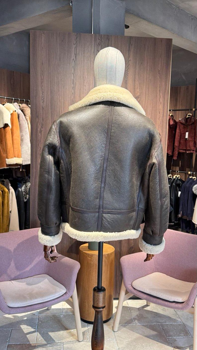 May include: A brown leather jacket with a cream-colored shearling collar and cuffs is displayed on a mannequin. The jacket has a classic design with a slightly distressed appearance. The back view shows the jacket's details, including seams and a warm, inviting aesthetic.