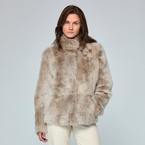 May include: A beige faux fur jacket with a high collar and long sleeves. The jacket features a thick, textured pile with subtle brown and grey undertones throughout the material. It is paired with plain off-white trousers, creating a neutral look.