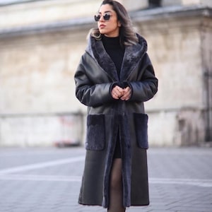 May include: A long, charcoal gray coat featuring a hood, long sleeves, and two large patch pockets. The coat is lined with dark gray faux fur, which is also visible along the front opening and the edges of the hood and pockets.