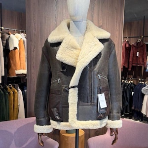 May include: A brown leather jacket with a cream-colored shearling lining and collar. The jacket features a classic design with a front closure, two pockets, and a tag. The jacket is displayed on a mannequin.