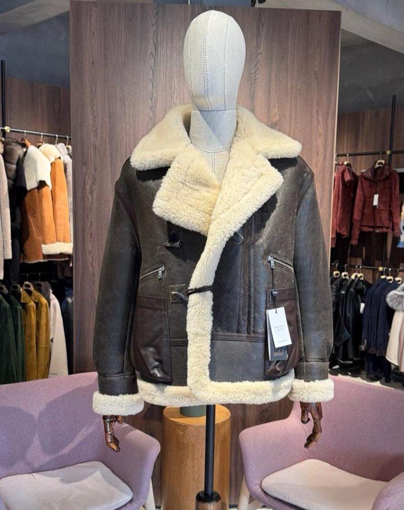 May include: A brown leather jacket with a cream-colored shearling lining and collar. The jacket features a classic design with a front closure, two pockets, and a tag. The jacket is displayed on a mannequin.