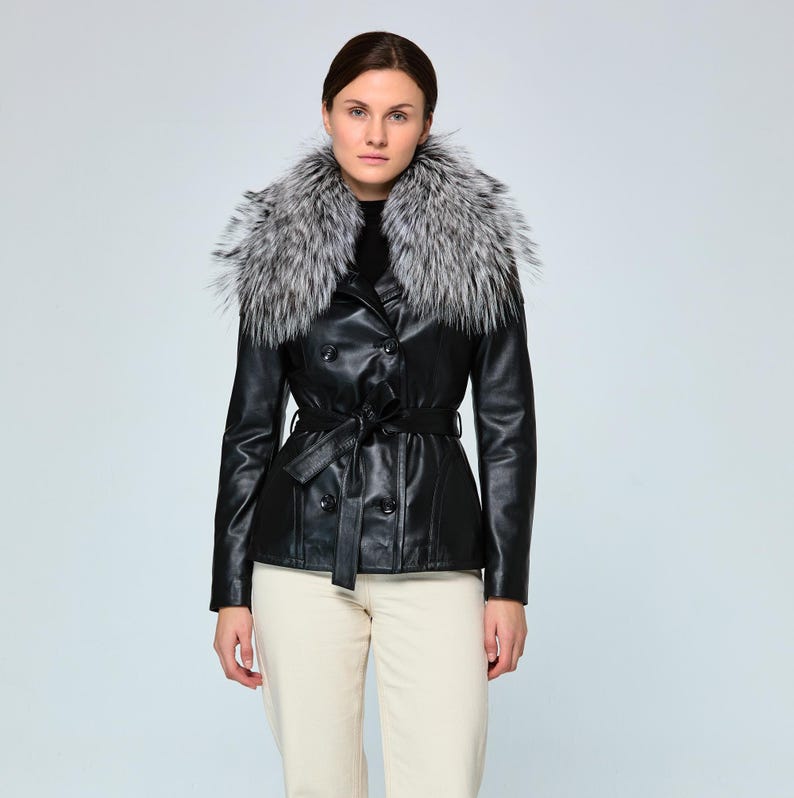 black leather jacket with a double-breasted design and a silver and gray fur collar. The jacket features a belted waist and is paired with cream-colored pants. The overall style is classic and elegant.