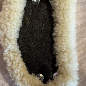 May include: Close-up of a sheepskin pad with a dark brown leather center. The pad is surrounded by a thick, cream-colored wool and secured with silver-colored rivets. The item is likely used for equestrian purposes.