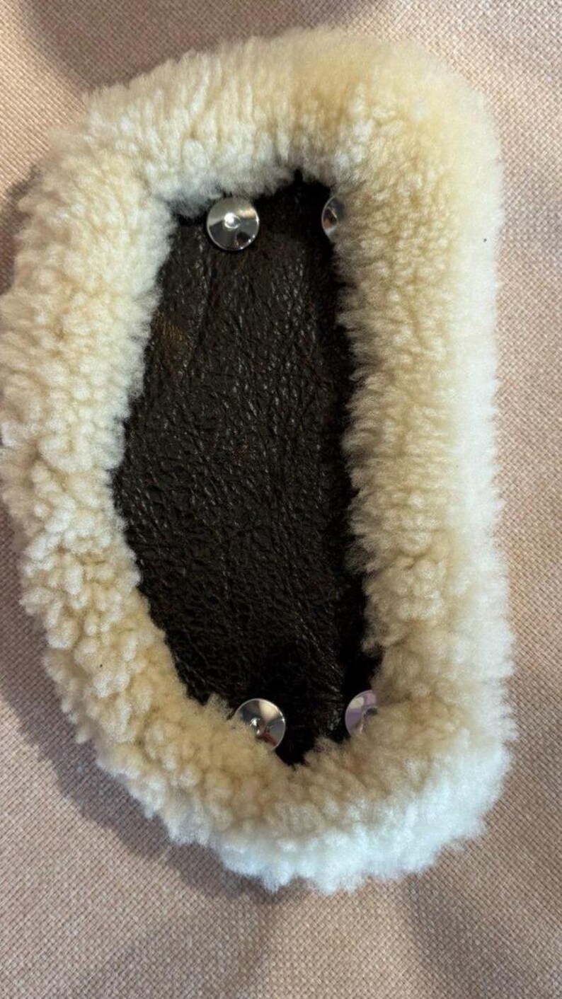May include: Close-up of a sheepskin pad with a dark brown leather center. The pad is surrounded by a thick, cream-colored wool and secured with silver-colored rivets. The item is likely used for equestrian purposes.