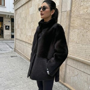 May include: A black, long-sleeved, high-neck jacket with a textured, fuzzy finish. The jacket features a front zipper closure and a small white rectangular tag with black text on the left sleeve cuff. The garment is worn over black leggings.