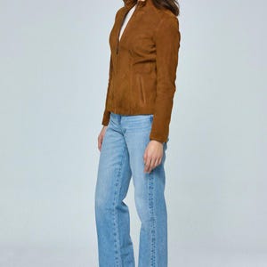 May include: A woman wearing a brown suede jacket with a zipper closure, light blue jeans, and tan heeled boots. The jacket has a high collar and a pocket on the left side. The jeans are a light wash and have a straight leg.