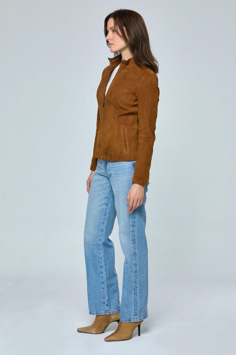 May include: A woman wearing a brown suede jacket with a zipper closure, light blue jeans, and tan heeled boots. The jacket has a high collar and a pocket on the left side. The jeans are a light wash and have a straight leg.