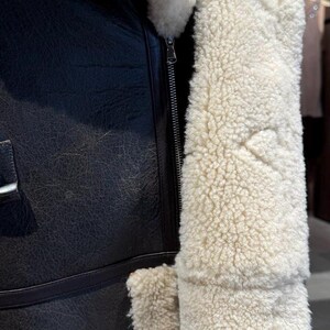 May include: Close-up of a black leather jacket with a cream-colored, fluffy shearling lining. The jacket features a silver zipper and a textured leather exterior. The shearling trim is visible along the collar and cuffs.