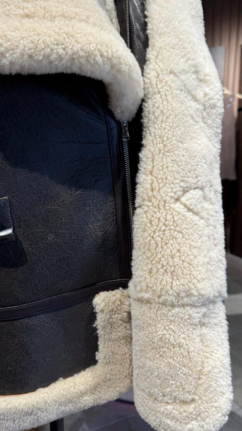 May include: Close-up of a black leather jacket with a cream-colored, fluffy shearling lining. The jacket features a silver zipper and a textured leather exterior. The shearling trim is visible along the collar and cuffs.