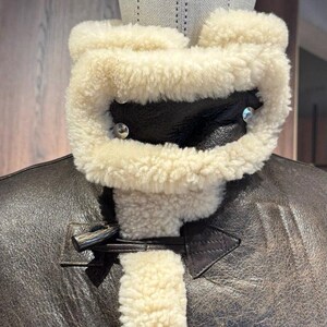 May include: Close-up of a brown leather jacket with a cream-colored shearling collar and lining. The jacket features a buckle closure and silver-toned hardware. The collar is secured with two silver rivets. The jacket is displayed on a mannequin.