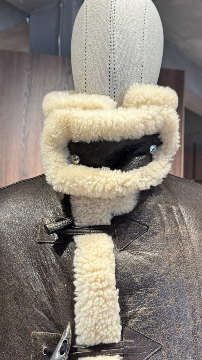 May include: Close-up of a brown leather jacket with a cream-colored shearling collar and lining. The jacket features a buckle closure and silver-toned hardware. The collar is secured with two silver rivets. The jacket is displayed on a mannequin.