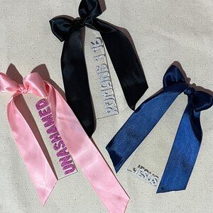 May include: Three decorative tags with satin ribbons. One tag has a pink ribbon and the word "UNASHAMED" in glitter. Another has a black ribbon with "ROMANS 1:16" in glitter. The third has a navy blue ribbon with "JESUS" in glitter.