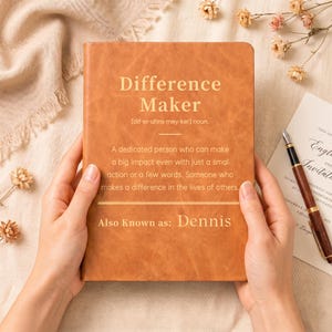 May include: A tan leather-bound journal featuring the gold-printed text Difference Maker, a phonetic pronunciation guide, a definition of the term, and the phrase Also Known as: Dennis, centred on the front cover in a clean, sans-serif font.