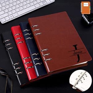 May include: A selection of ring-bound notebooks in black, red, navy, and brown. Each notebook has a personalised cover with the name "Jennie". The notebooks are displayed on a dark surface, with a keyboard visible in the background.