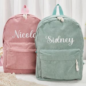 May include: Two corduroy backpacks, one pink and one teal, are displayed. Each backpack has a front pocket and a top handle. The pink backpack has the name "Nicole" embroidered in white, while the teal backpack has "Sidney" embroidered in white.