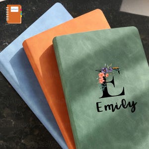 May include: Three notebooks stacked on top of each other. The top notebook is green with a floral design and the letter "E" with the name "Emily" written below it. The middle notebook is orange and the bottom notebook is blue.