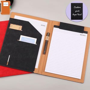 Puede incluir: Una carpeta de cuero negro con un borde rojo y una solapa marrón. La carpeta tiene un bloc de notas, un portaplumas y un bolsillo para tarjetas de visita. La carpeta tiene el texto "MEMO" impreso en la parte delantera.