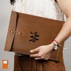 May include: A brown leather portfolio with a flap closure and a button clasp. The portfolio features the name "Williams" in a stylized font. The portfolio is held by a person.
