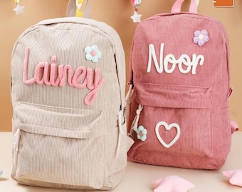 May include: Two corduroy backpacks, one beige and one pink. The beige backpack has the name "Lainey" in pink script. The pink backpack has the name "Noor" in white script, a white heart, and a flower. Both have a front pocket.
