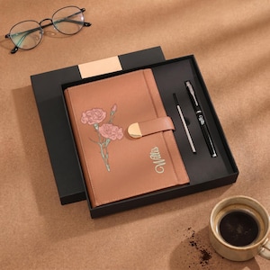 May include: A tan leather-textured notebook with a floral illustration of two pink carnations and the name Willis printed in script. The notebook features a gold-toned magnetic closure and is presented in a black gift box with a black pen.