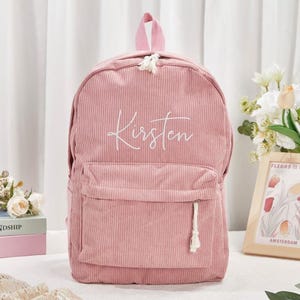 May include: A pink corduroy backpack with the name "Kirsten" embroidered in white script. The backpack has a front pocket, a top handle, and a tassel zipper pull. The bag is a soft pink color.