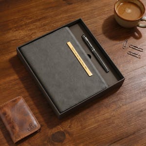 May include: A gift set comprising a grey journal with a gold bar inscribed with "Don't Let the Hard Days Win", a pen, and a leather wallet. A cup of coffee and paper clips are also visible.