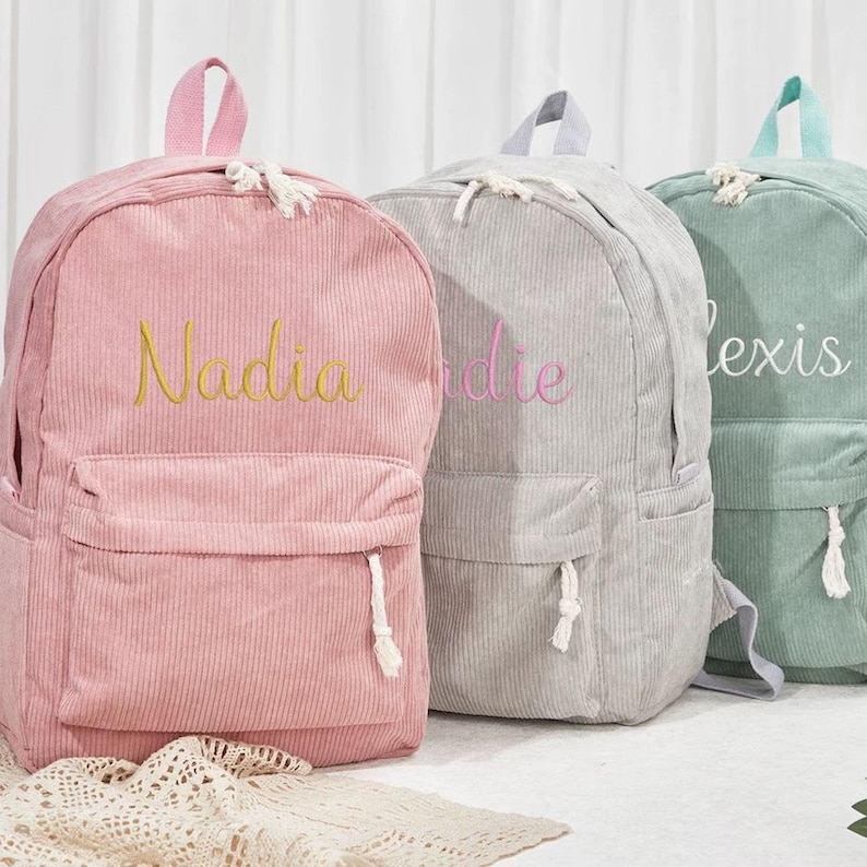 May include: Three corduroy backpacks in pink, gray, and green. Each backpack has a front pocket, a top handle, and adjustable straps. The pink backpack has the name "Nadia" embroidered in yellow, the gray backpack has "die" in pink, and the green backpack has "lexis" in white.