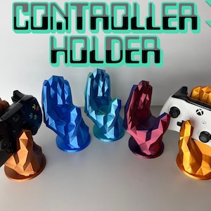 Könnte beinhalten: Fünf 3D-gedruckte, handförmige Halter für Spielecontroller. Die Halter sind in Blau, Türkis, Rot und Gold. Die Halter halten zwei verschiedene Spielecontroller, einen schwarzen und einen weißen.