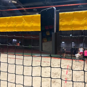 Könnte beinhalten: Ein schwarzes Smartphone wird in einer schwarzen Halterung an einem gelben Netz befestigt. Das Netz ist Teil eines Volleyballfeldes. Der Bildschirm des Telefons ist sichtbar, mit einem Sandplatz und Personen im Hintergrund.
