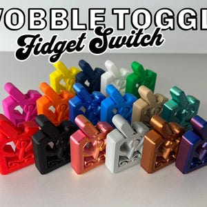 Può includere: Una collezione di 15 interruttori a levetta oscillanti per fidget di diversi colori. Gli interruttori sono in plastica e presentano una varietà di colori, tra cui rosso, blu, verde, giallo e nero. Gli interruttori hanno tutti la forma di un interruttore a levetta e hanno una piccola manopola rotonda sulla parte superiore. Il testo "WOBBLE TOGGLE Fidget Switch" è visualizzato nella parte superiore dell'immagine.