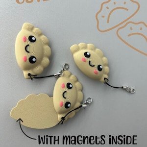 Könnte beinhalten: Drei beige, knödelartige Magnete mit lächelnden Gesichtern und schwarzen Kordeln. Der Text "CUTE DUMPLINGS" steht über den Magneten, und "WITH MAGNETS INSIDE" steht darunter. Die Knödel haben rosige Wangen und schwarze Augen.