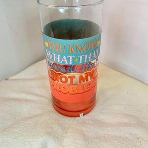 May include: A clear glass tumbler with a teal band and orange stripes. The tumbler has the text "YOU KNOW WHAT THAT sounds like? NOT MY PROBLEM" in a playful font. The glass is cylindrical and appears to be empty.