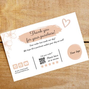 Op de afbeelding: Een bedankkaart met een perzikkleurige achtergrond en een hart in de rechterbovenhoek. De kaart zegt "Bedankt voor uw aankoop!" en bevat een QR-code, vijf sterren en de tekst "Laat een beoordeling achter".