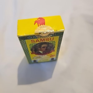 Może przedstawiać: Żółte pudełko z napisem "SAMSU" w zielonych i złotych literach. Pudełko zawiera wizerunek mężczyzny w garniturze i okularach. Na górze pudełka znajduje się czerwone logo konia.