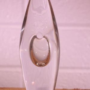 May include: A clear glass vase with a teardrop shape and a circular bubble in the center. The vase is set against a pinkish-white brick wall and a wooden surface. The design is minimalist and modern.