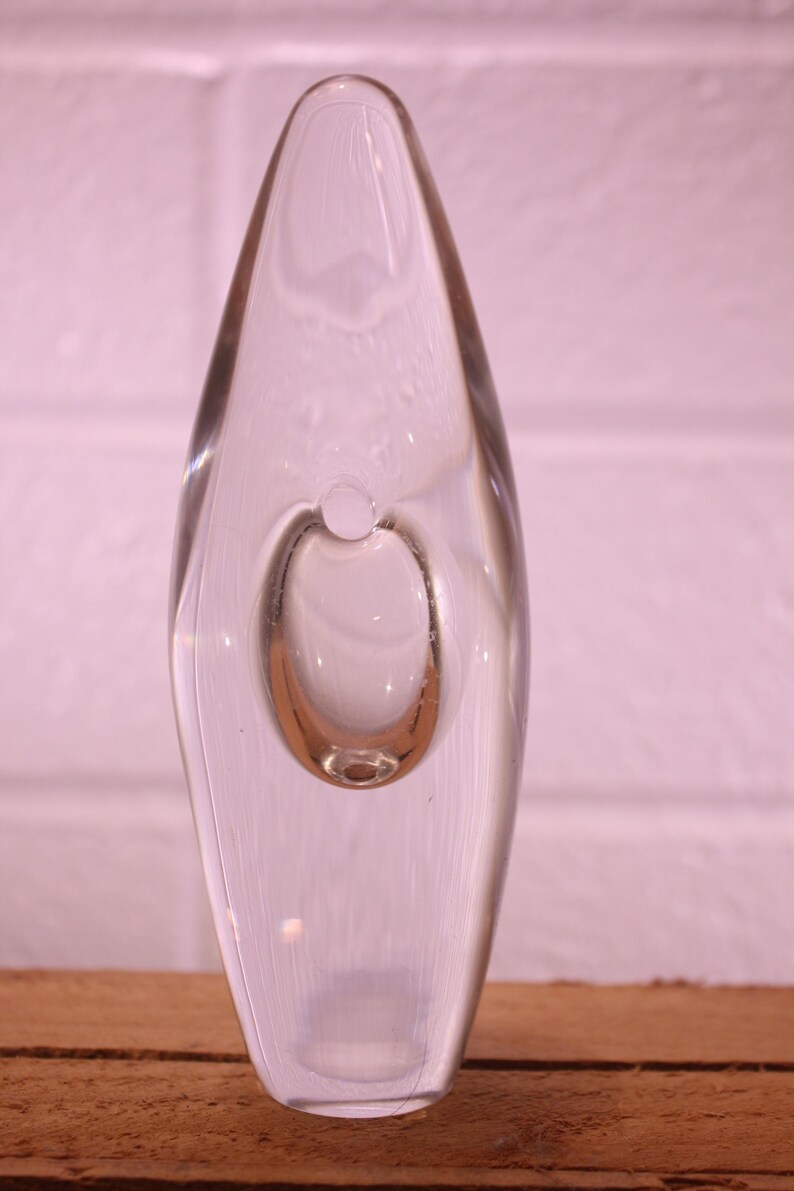 May include: A clear glass vase with a teardrop shape and a circular bubble in the center. The vase is set against a pinkish-white brick wall and a wooden surface. The design is minimalist and modern.